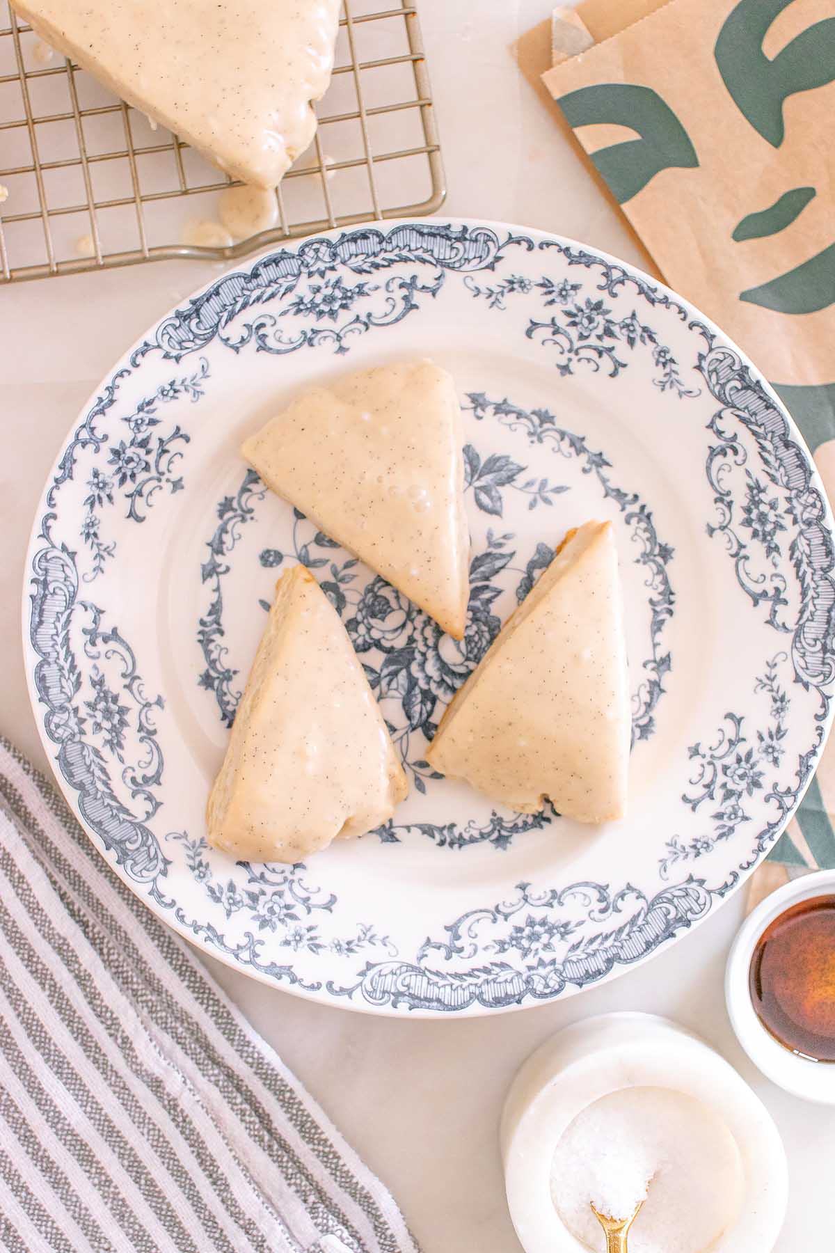 Three glazed Starbucks Copycat Vanilla Bean Scones rest on a blue and white floral plate, with baking items and a striped towel nearby.
