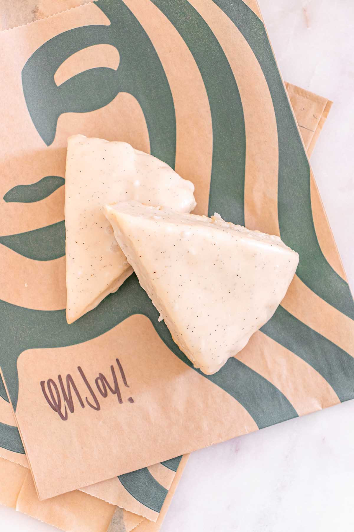 Two Starbucks Copycat Vanilla Bean Scones with icing rest on Starbucks-branded brown paper bags, one featuring the word "enjoy!".