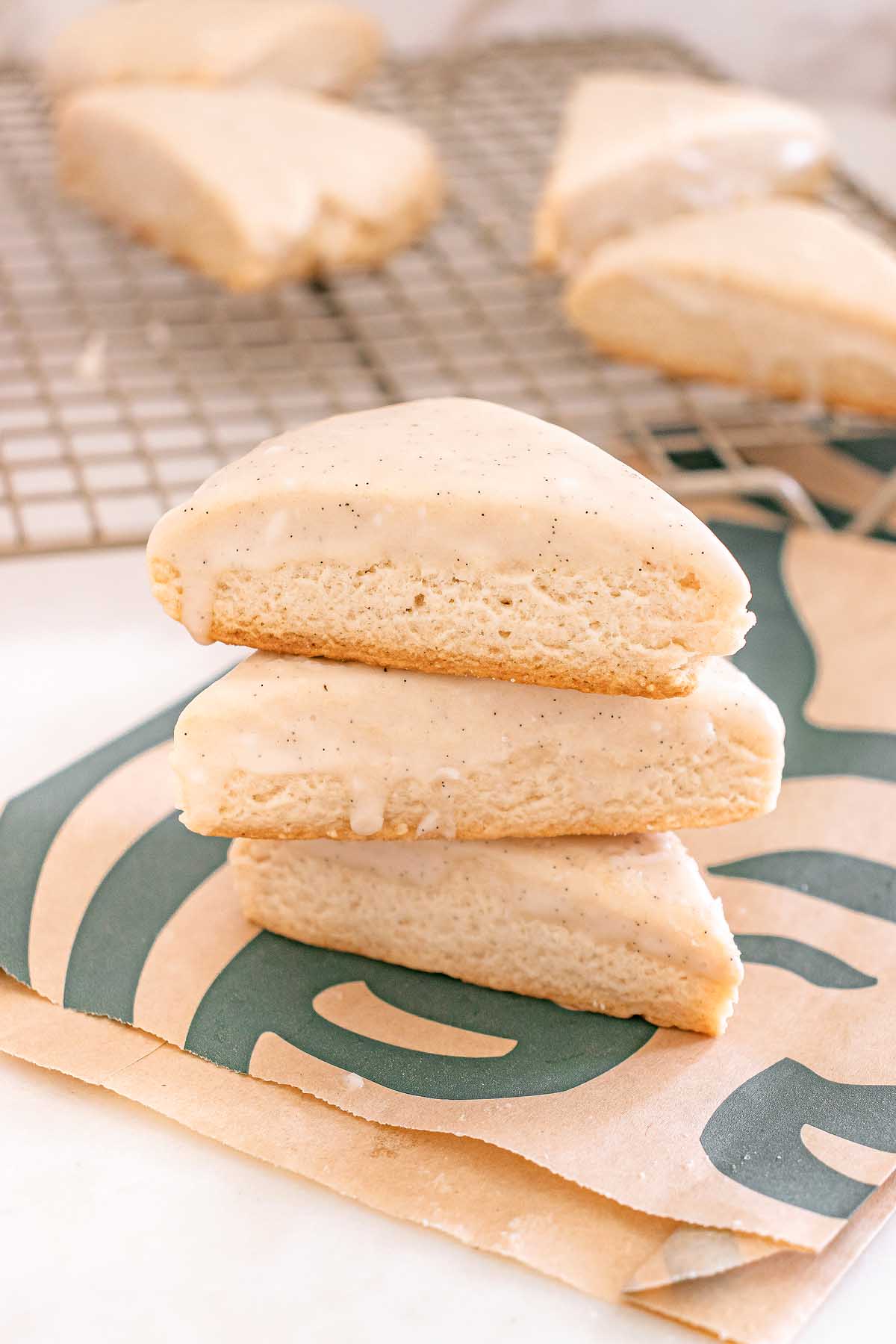 Three Starbucks Copycat Vanilla Bean Scones stacked on a Starbucks bag, with more scones cooling on a rack in the background.