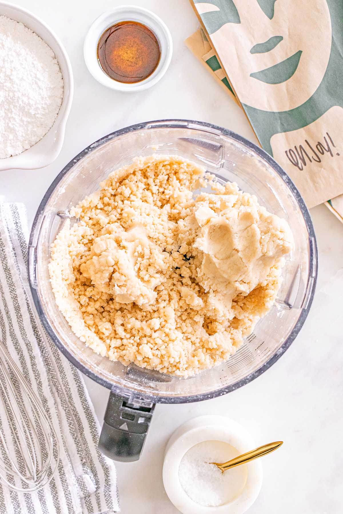 Overhead view of a food processor with cookie dough for Starbucks Copycat Vanilla Bean Scones, sugar, salt, vanilla, and a striped towel on a white surface.