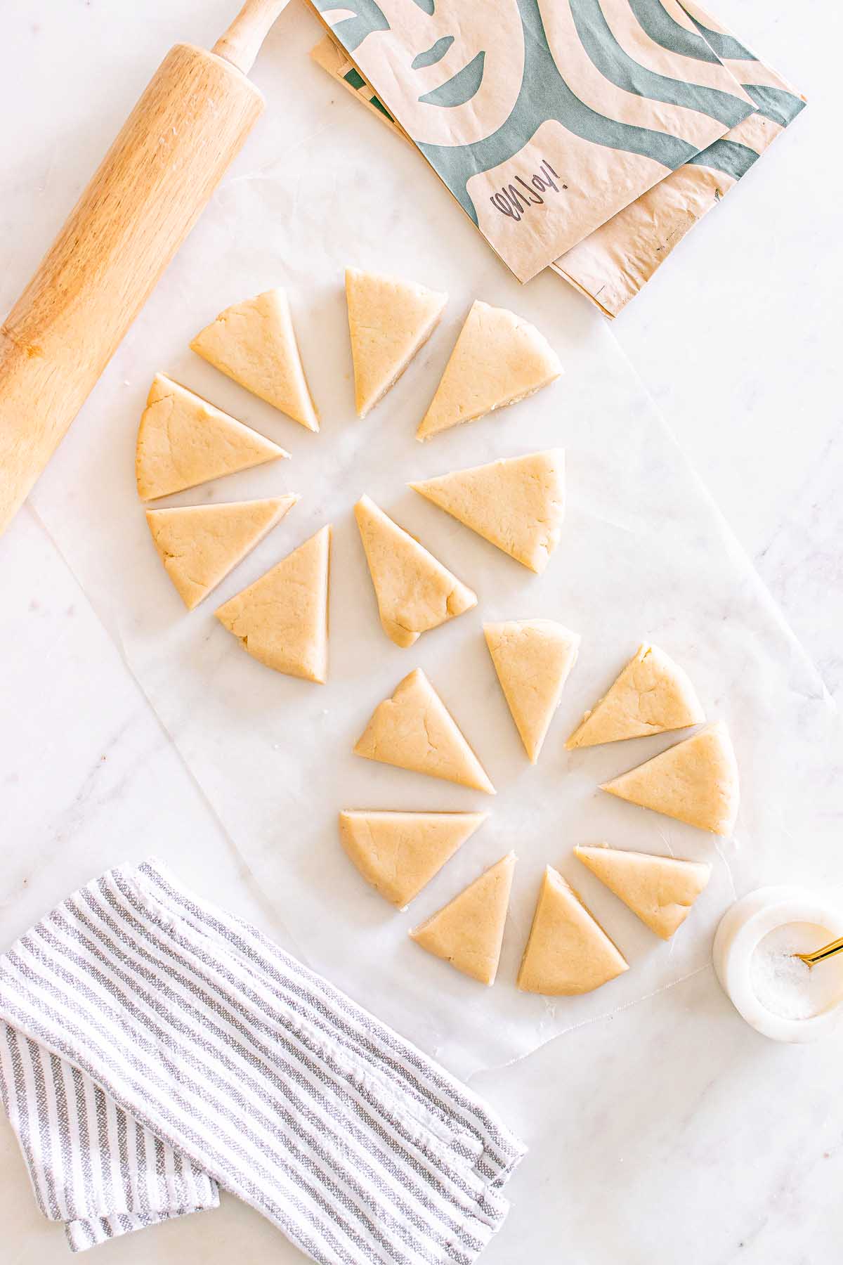 Two circles of dough cut into wedges on parchment, ready to become Starbucks Copycat Vanilla Bean Scones, with a rolling pin, striped towel, and paper bag nearby.