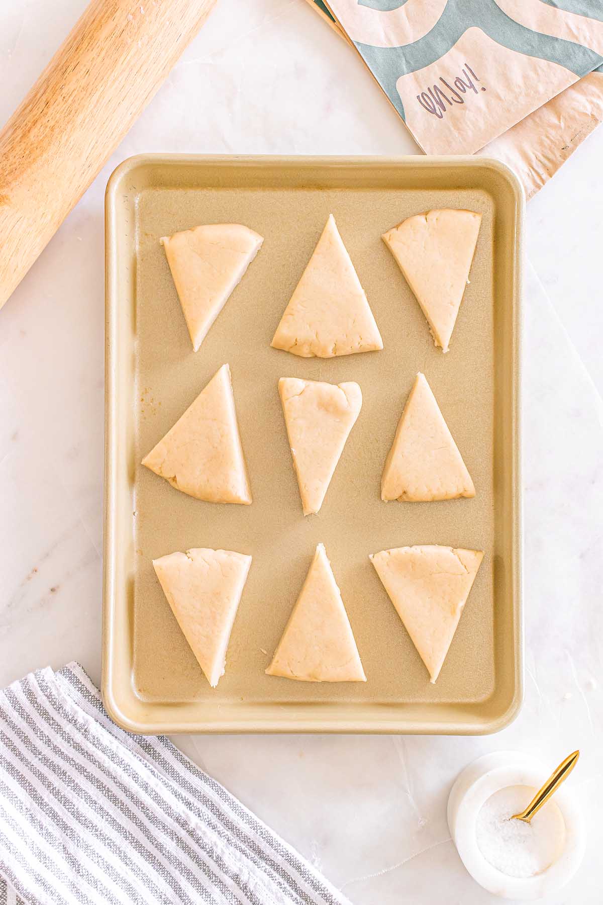 Nine unbaked triangular Starbucks Copycat Vanilla Bean Scones on a baking sheet, ready to bake, with baking tools nearby.