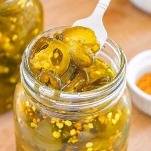 A fork lifts pickled jalapeños, also known as cowboy candy, from a glass jar next to a small bowl of spices on a wooden surface.