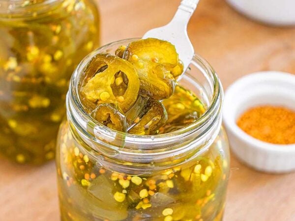 A fork lifts pickled jalapeños, also known as cowboy candy, from a glass jar next to a small bowl of spices on a wooden surface.
