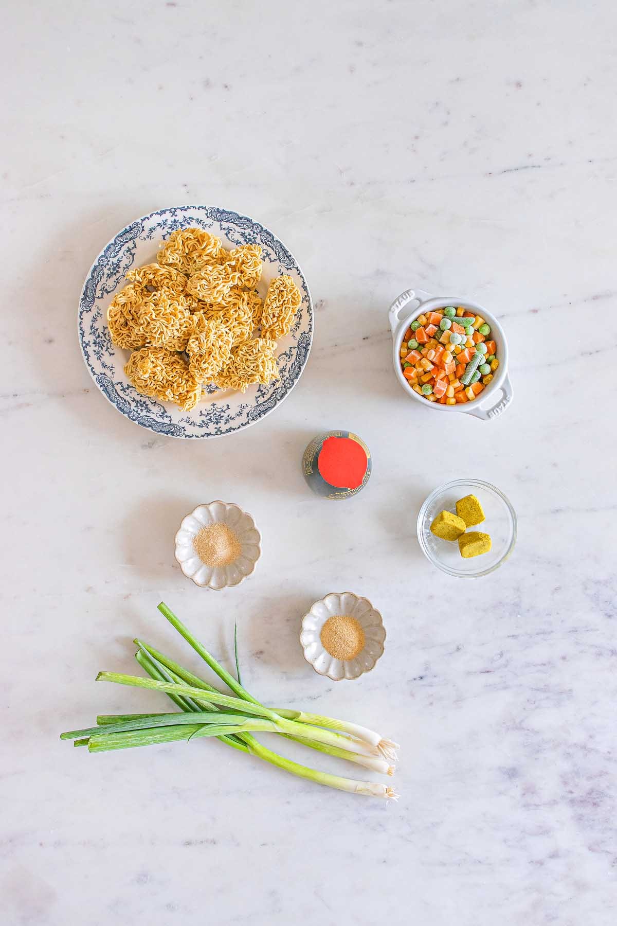 Ramen noodles, mixed vegetables, green onions, bouillon cubes, soy sauce, and seasonings arranged for a tasty mason jar cup of noodles on a white surface.