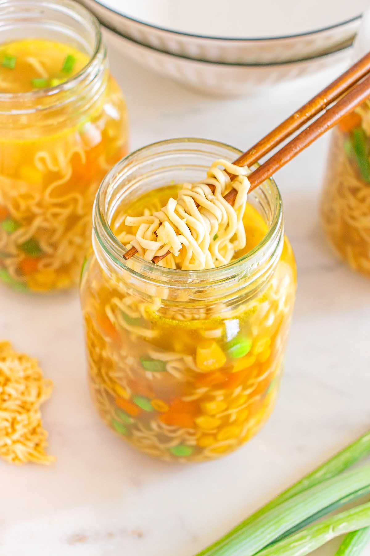 Chopsticks lifting noodles from a mason jar cup of noodles filled with vegetable ramen soup on a white surface.