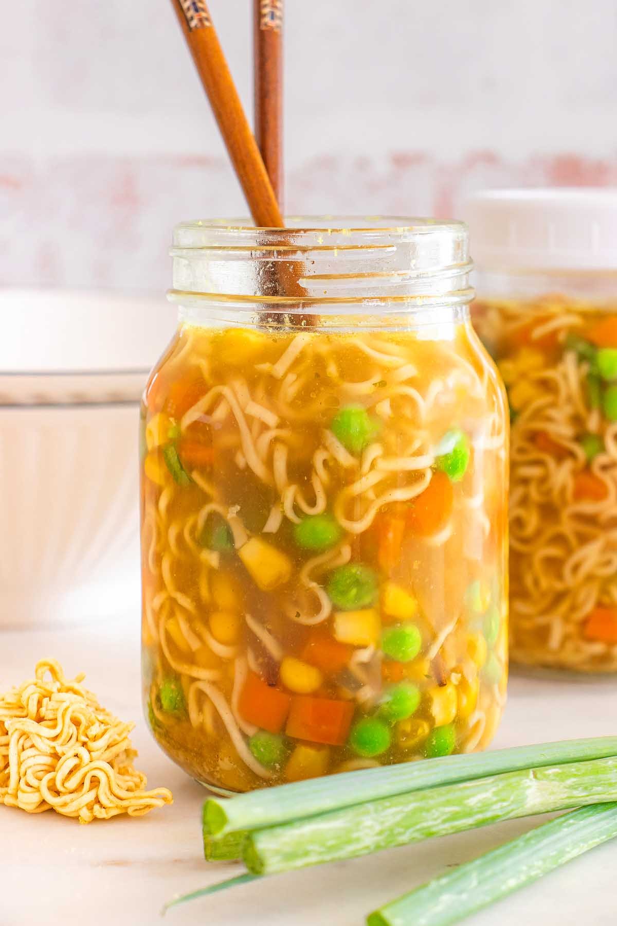 A mason jar cup of noodles filled with soup, mixed vegetables, and chopsticks, with scallions and dry noodles nearby.