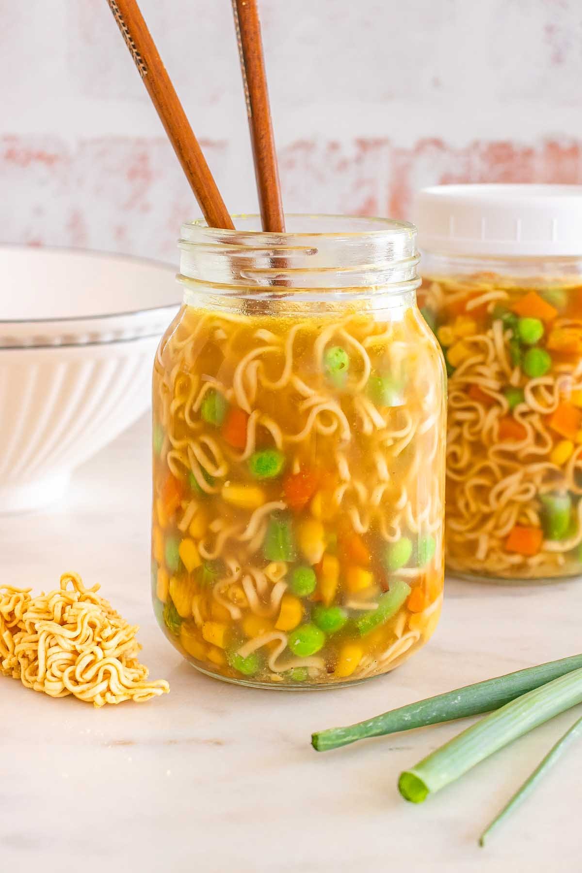 A mason jar cup of noodles filled with vegetables and chopsticks, with another jar and fresh ingredients in the background.