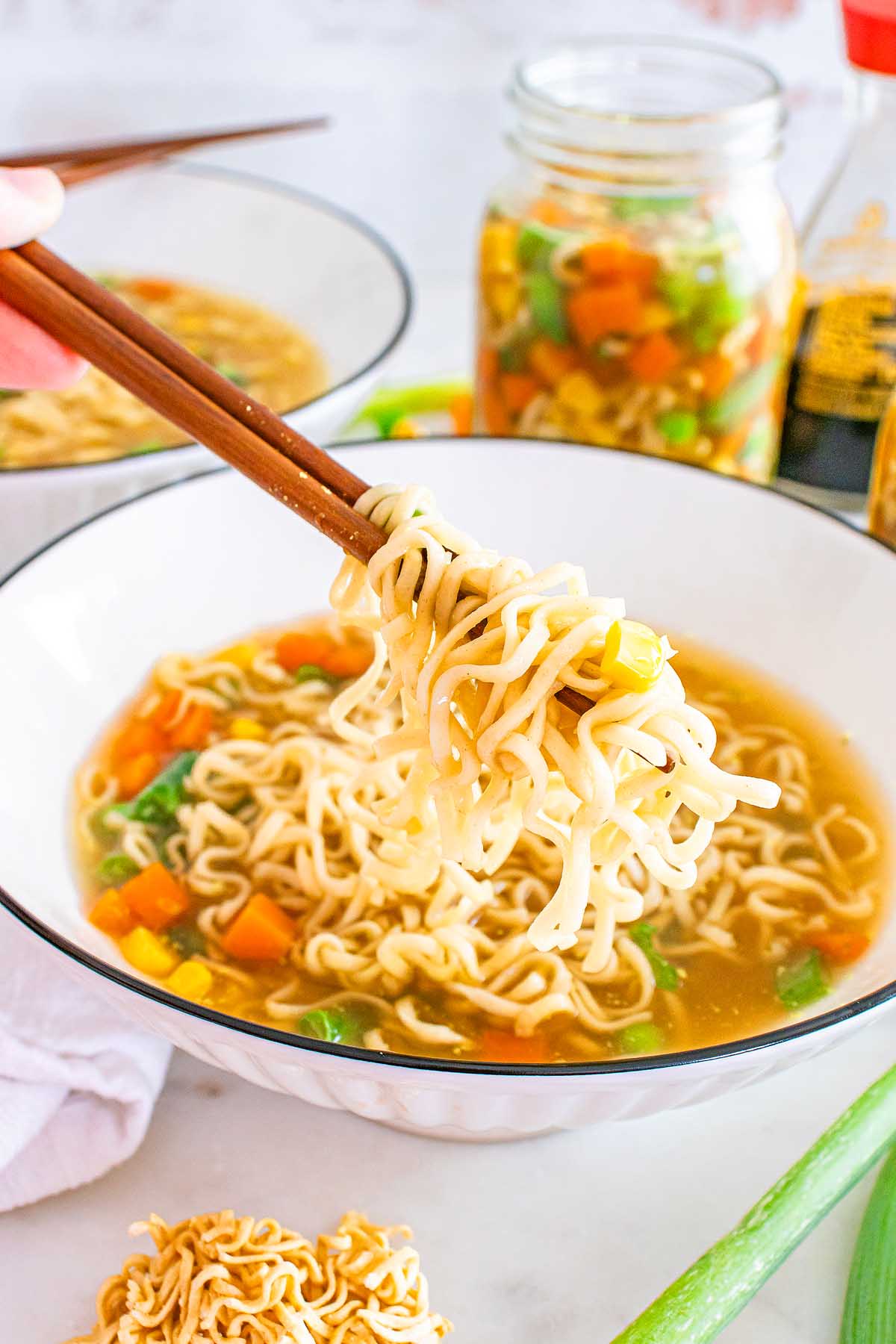 Chopsticks holding ramen noodles over a bowl of soup with vegetables and broth, inspired by the convenience of a mason jar cup of noodles.