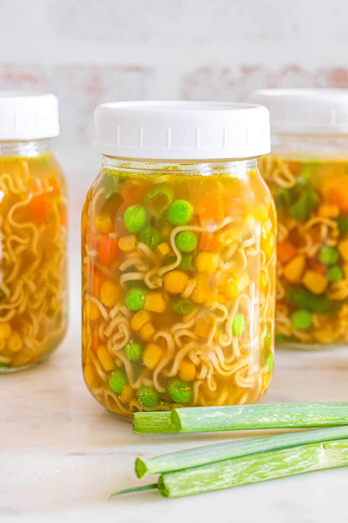 Three mason jar cups of vegetable noodle soup with peas, corn, and carrots sit next to green onions on a white surface.