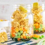 Two mason jar cup of noodles filled with ramen, mixed vegetables, and pickles sit on a striped surface, with soy sauce in the background.