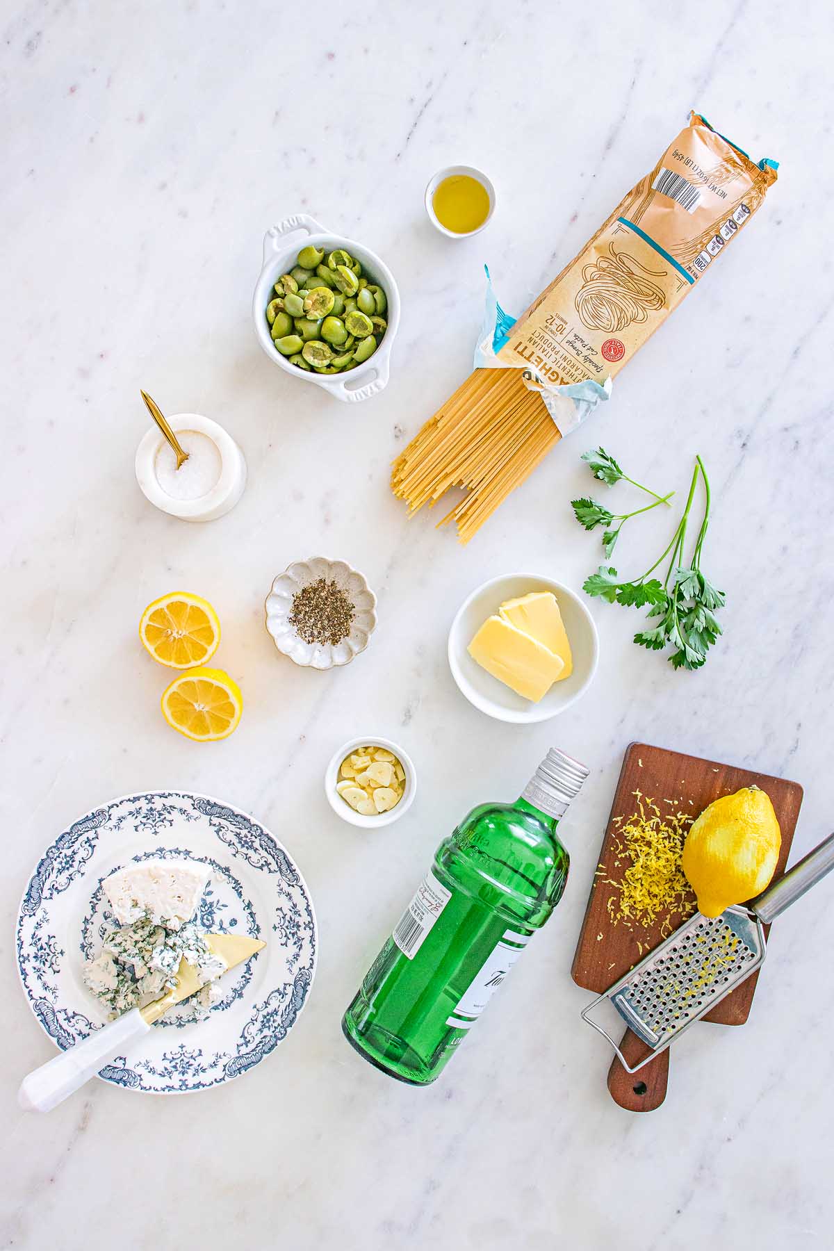 Flat lay of pasta, olives, lemon, butter, parsley, olive oil, cheese, gin, and seasonings on a marble surface—perfect ingredients for a dirty martini pasta.