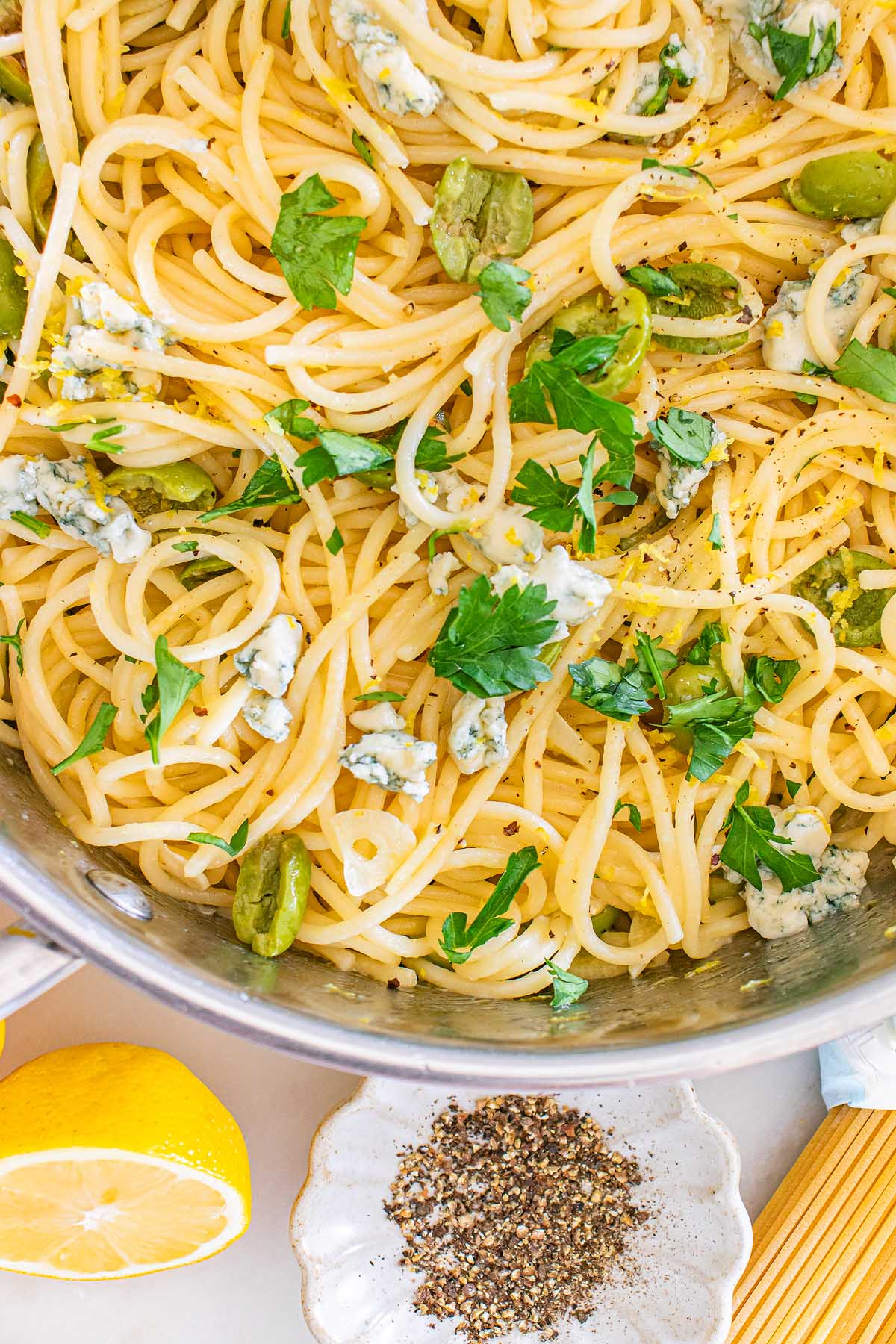 Close-up of dirty martini pasta featuring spaghetti with green olives, parsley, blue cheese, and black pepper in a pot, with lemon and pepper nearby.