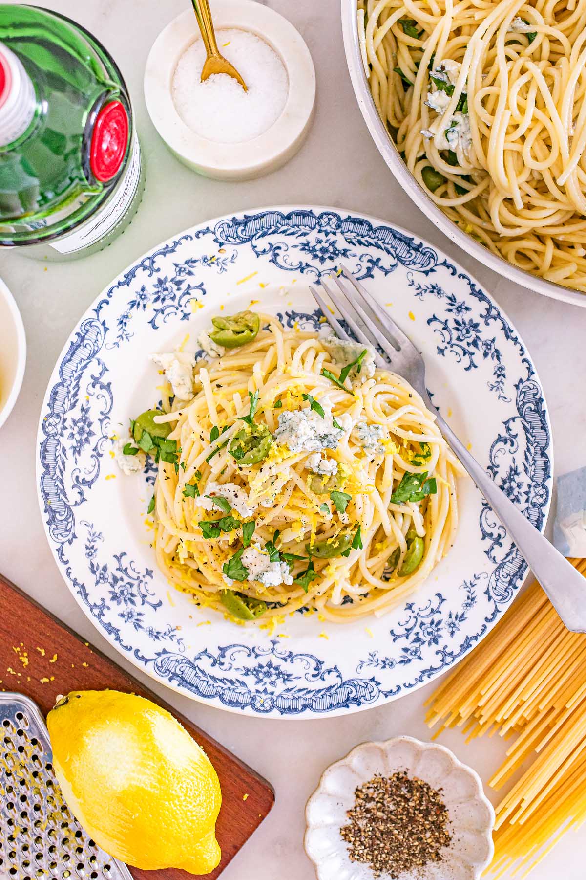 Plate of spaghetti with greens, cheese, and lemon zest on a patterned plate, surrounded by pasta ingredients—perfect for pairing with a dirty martini pasta twist.