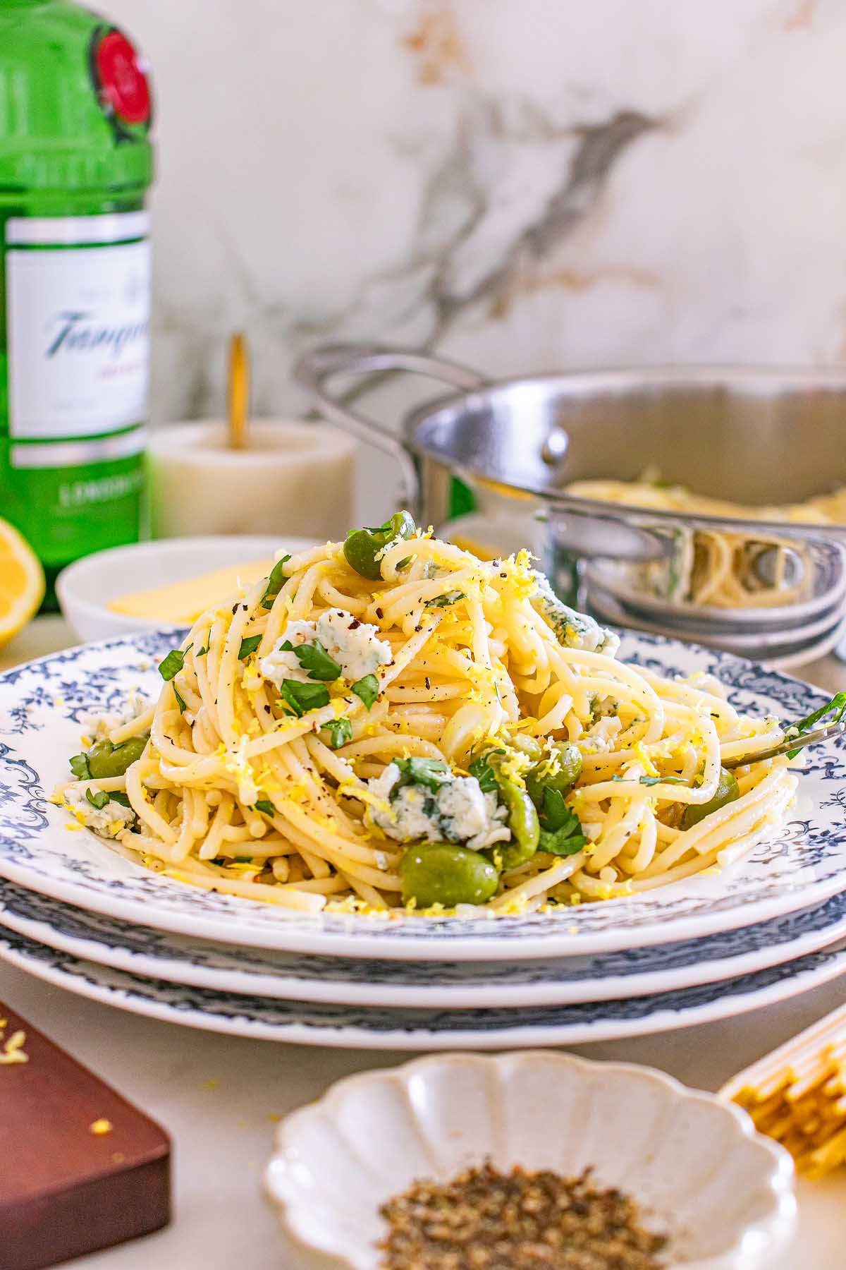 Spaghetti pasta with green olives and herbs on a plate, inspired by dirty martini pasta, with a bottle and pot in the background.