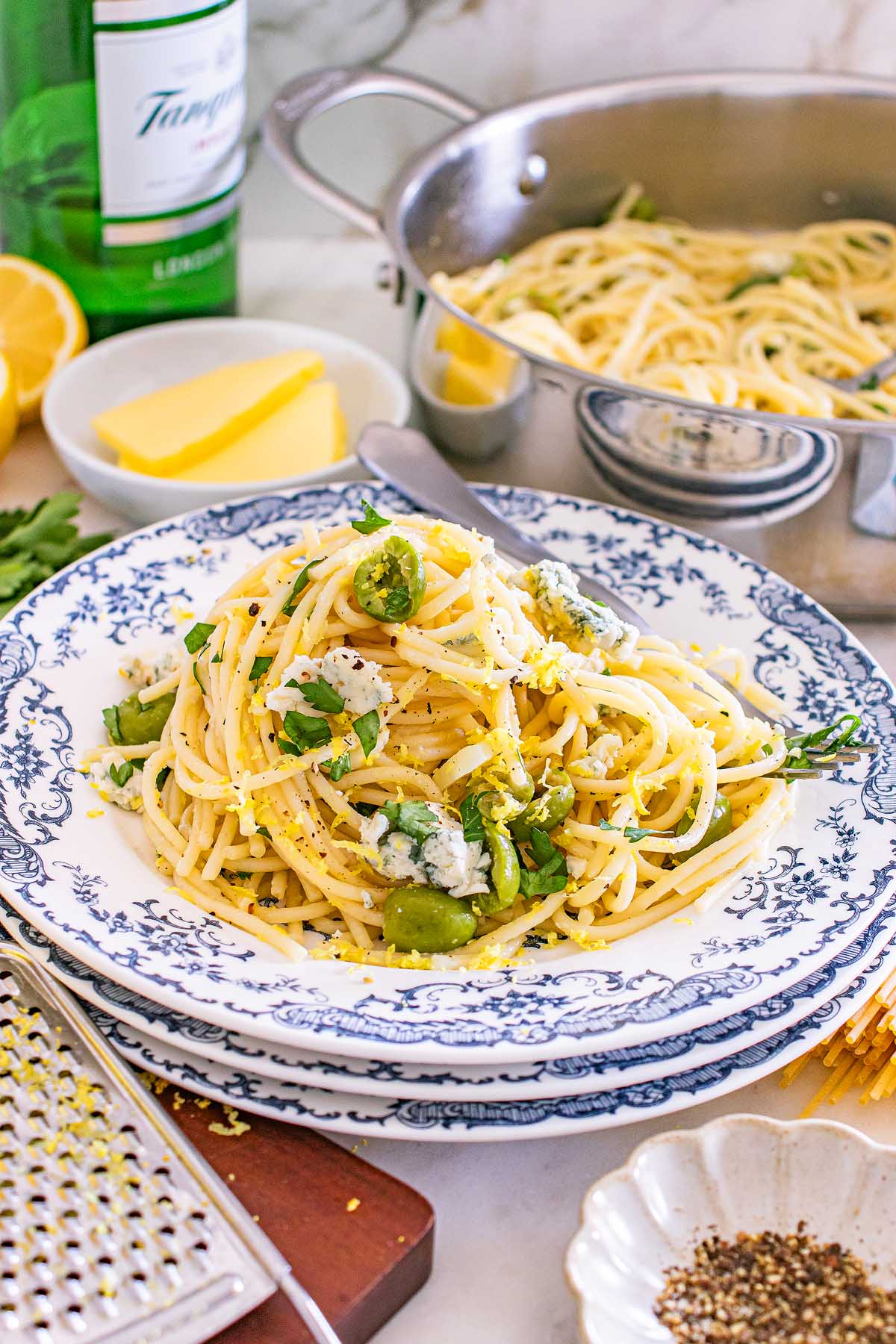 A plate of spaghetti with green vegetables, herbs, cheese, and butter, inspired by the bold flavors of dirty martini pasta, with a pot of pasta in the background.