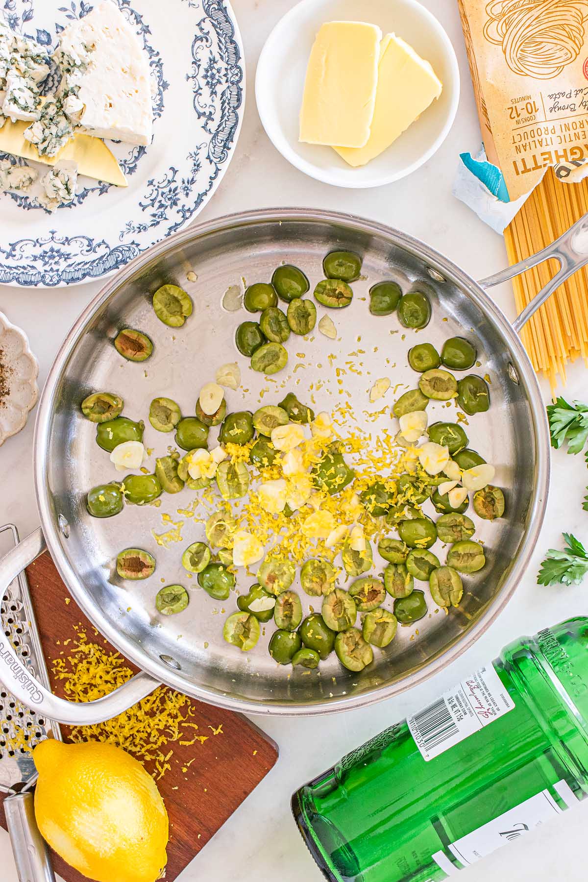 Sliced green olives, garlic, and lemon zest sizzle in a stainless steel pan—a perfect base for dirty martini pasta, with fresh ingredients artfully arranged around it.