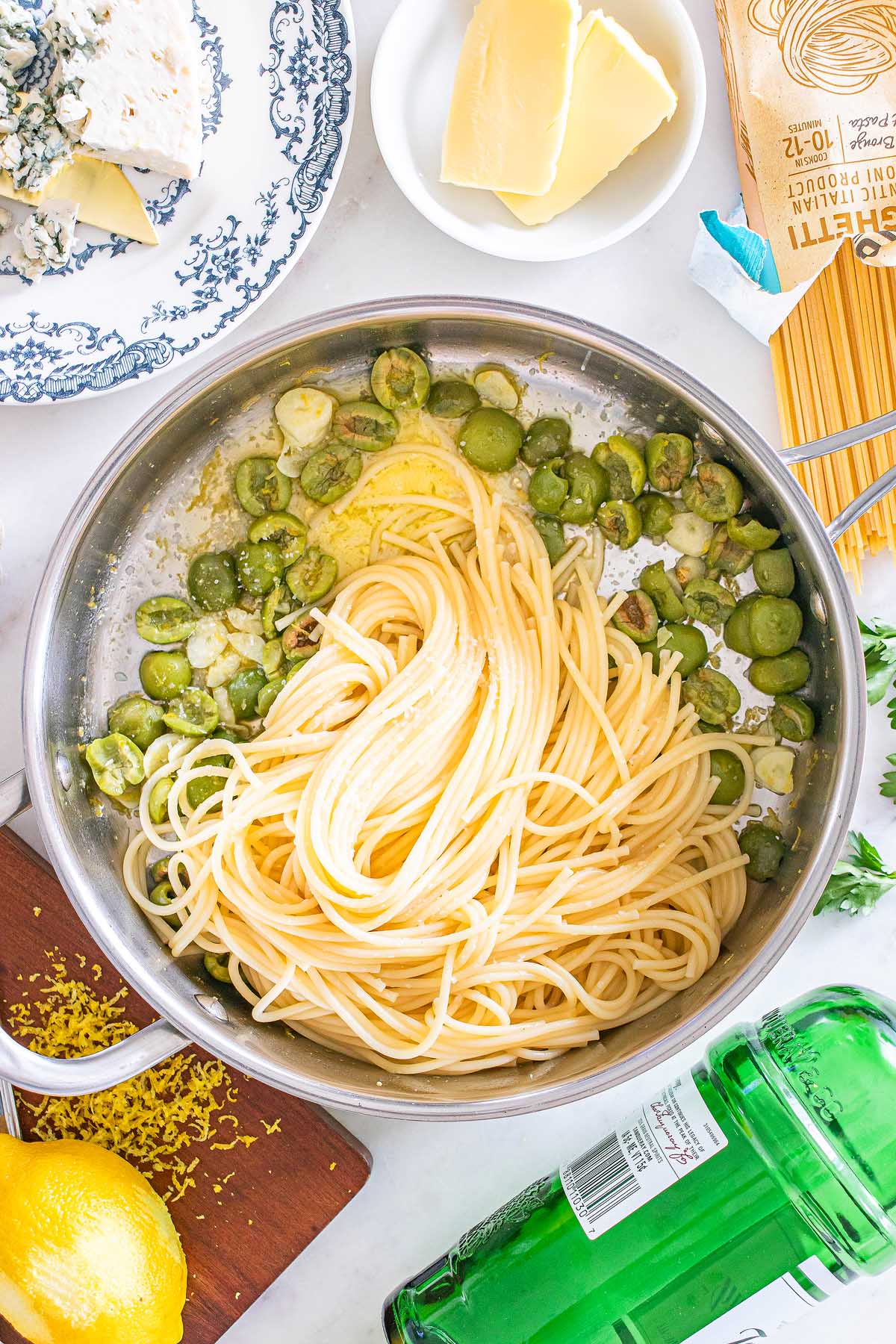 Cooked spaghetti in a pan with butter and green vegetables, inspired by dirty martini pasta, surrounded by ingredients on a white surface.