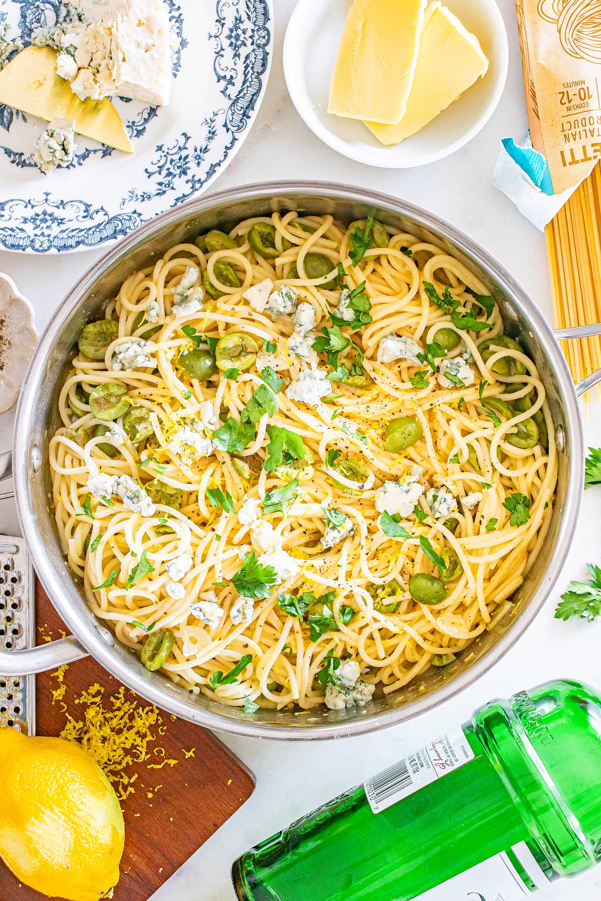 A pot of dirty martini pasta with green olives, herbs, and cheese, surrounded by lemon zest, butter, and assorted kitchen items.