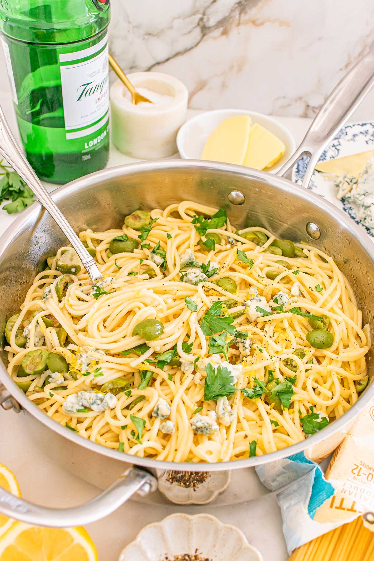 A pan of dirty martini pasta with green olives, parsley, and cheese sits on a kitchen counter, surrounded by fresh ingredients.