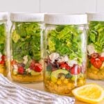 Four mason jars filled with vibrant Mason Jar Mediterranean Salad layers sit on the counter, accompanied by a lemon wedge and a striped towel.