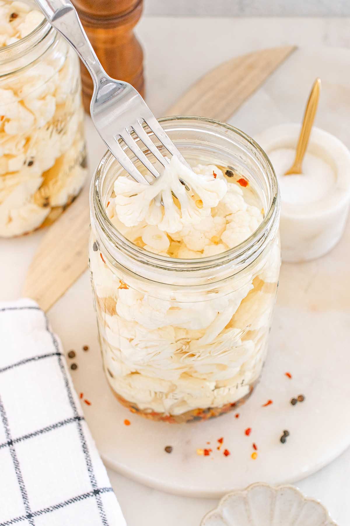 A fork lifting tangy pickled cauliflower from a glass jar, surrounded by vibrant spices on a marble surface.
