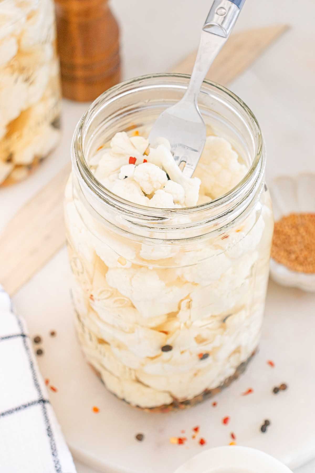 A fork in a jar of pickled cauliflower with spices sits on a white surface, showcasing the tangy flavor and vibrant colors of pickled cauliflower.