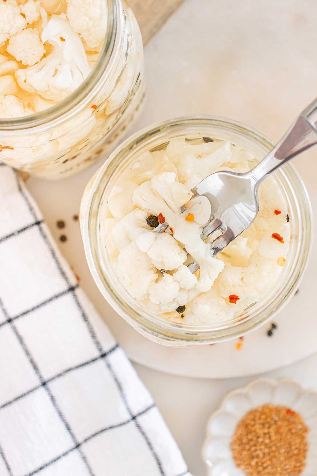 A fork holds tangy pickled cauliflower from a glass jar, with aromatic spices and a striped towel nearby.