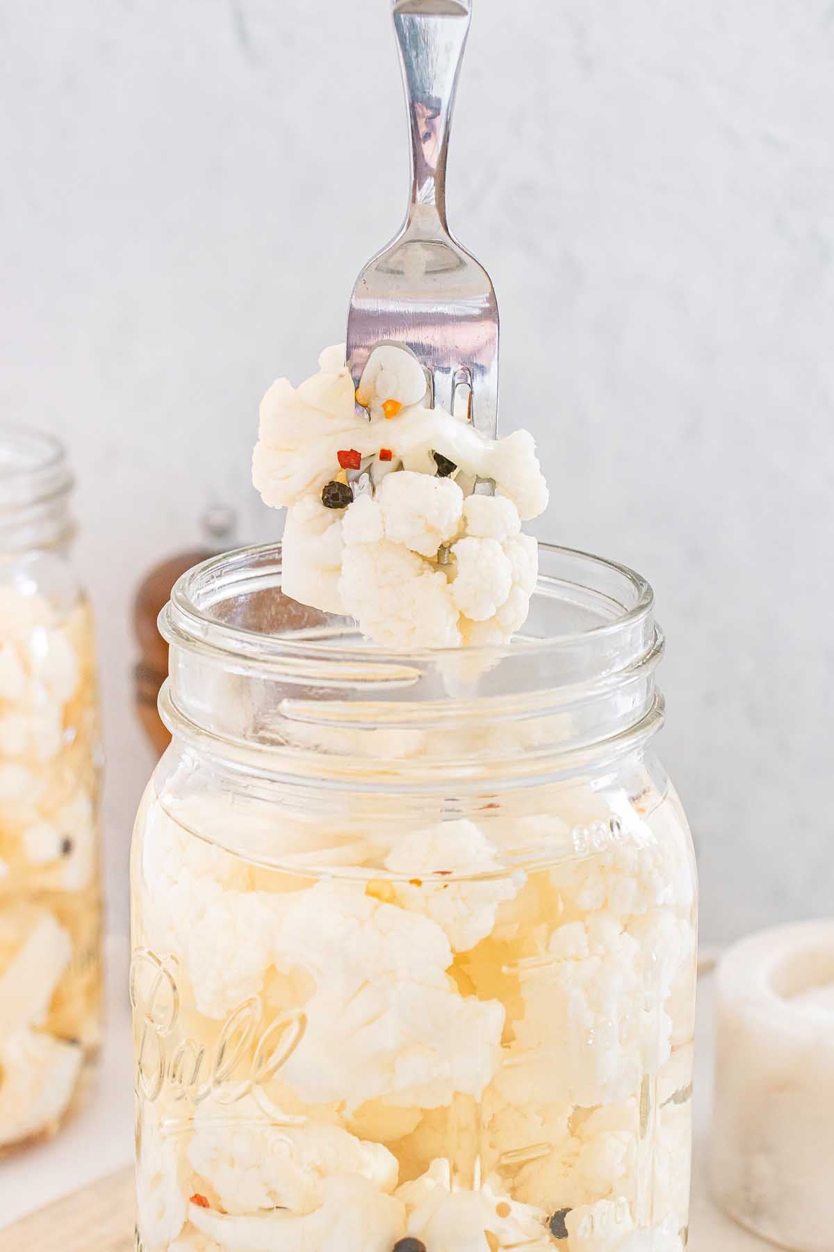 A fork holds pickled cauliflower above a glass jar filled with flavorful pickled cauliflower and spices.