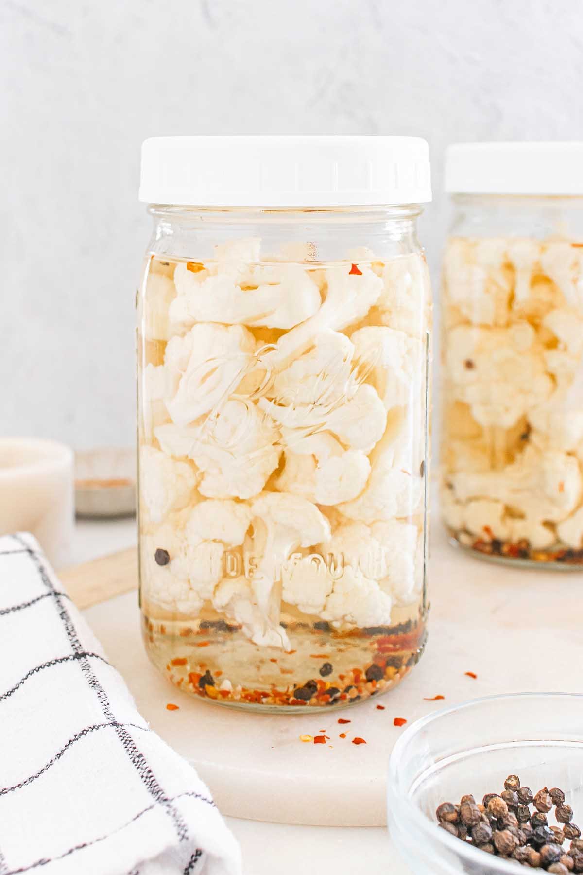 A jar of pickled cauliflower florets with spices sits on a white surface next to a striped towel, ready to enjoy as a tangy snack.