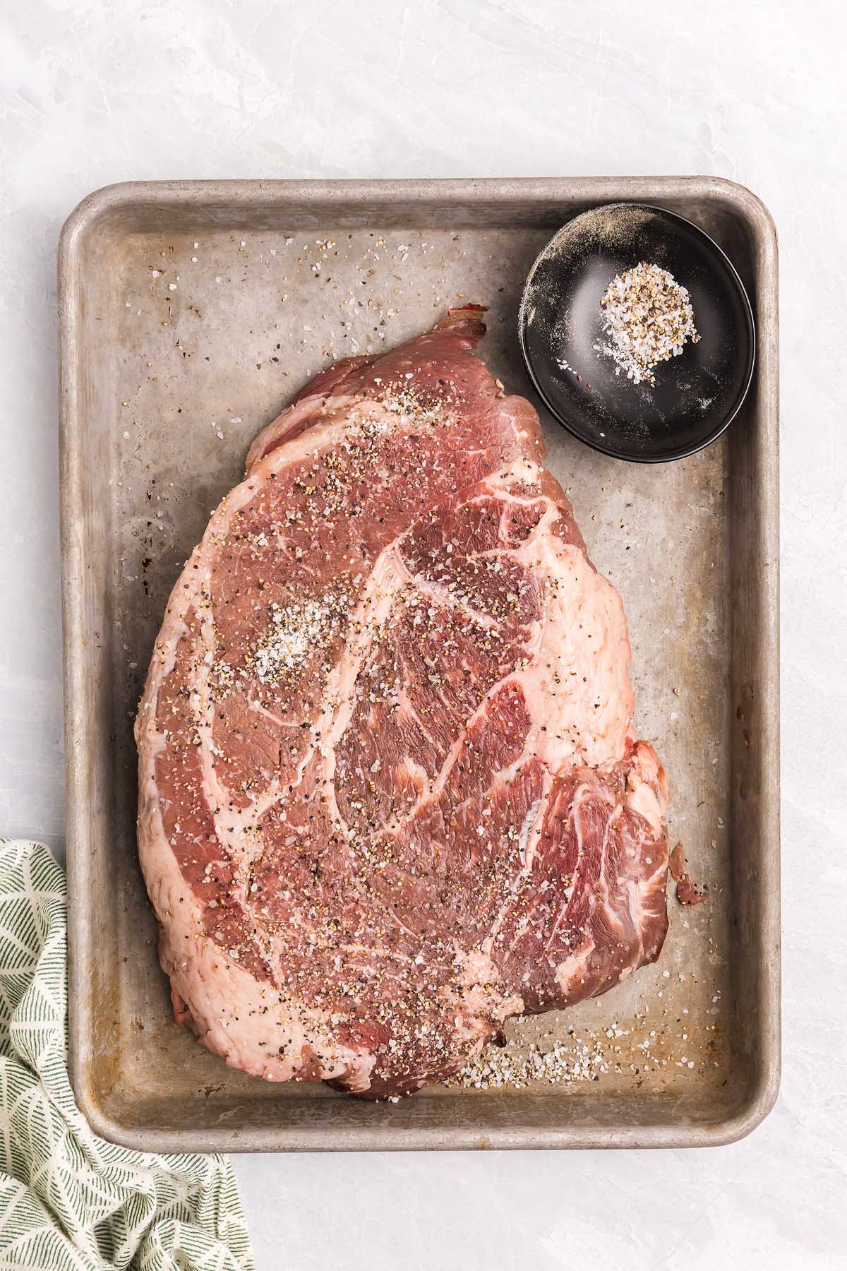 Raw ribeye steak seasoned with salt and pepper on a metal baking sheet, alongside extra seasoning in a small bowl&mdash;perfect to pair with pasta alla Genovese for a hearty meal.