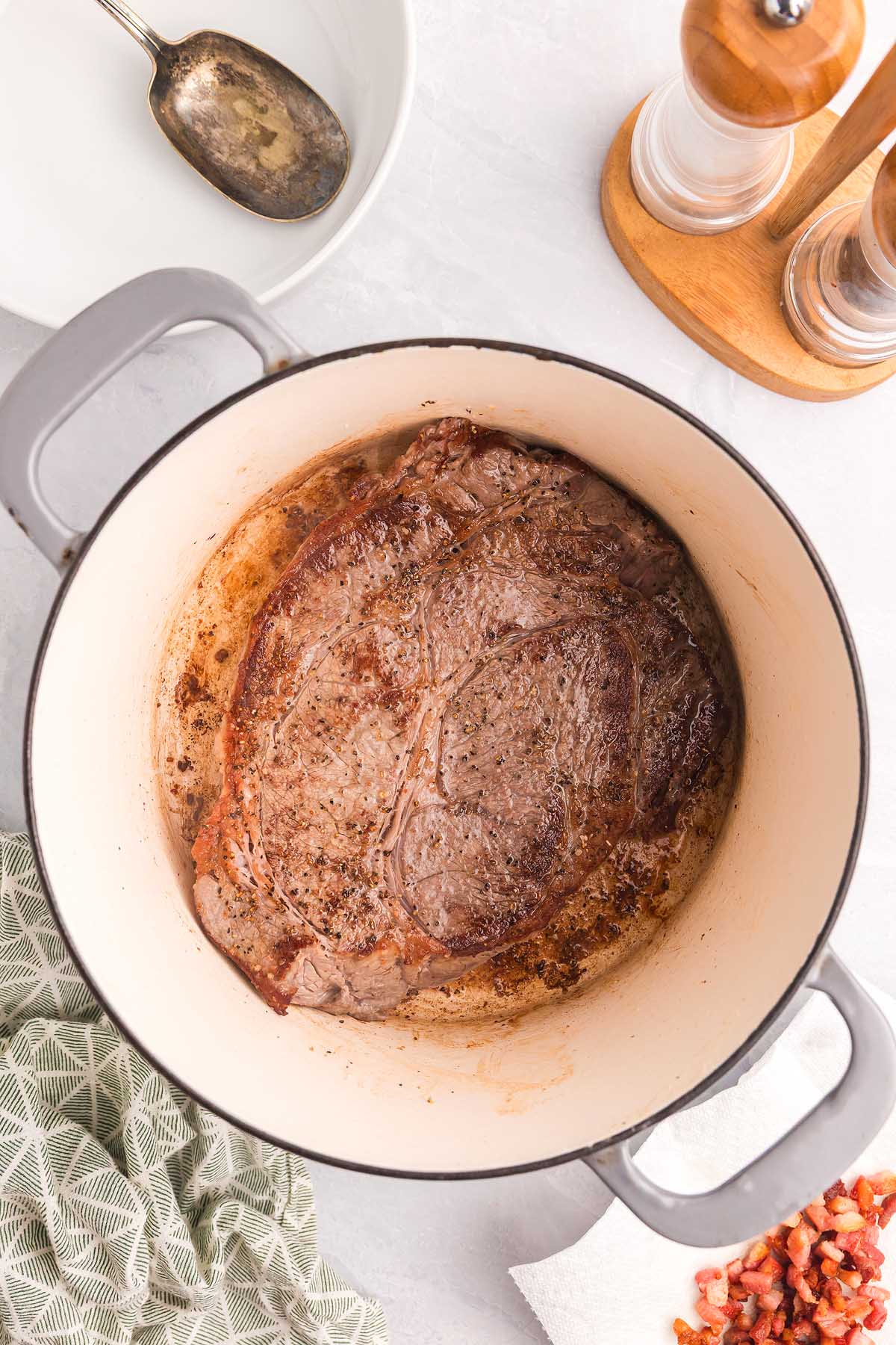 A seared steak in a white Dutch oven, surrounded by kitchen utensils, seasonings, and hints of pasta alla Genovese ingredients on a countertop.