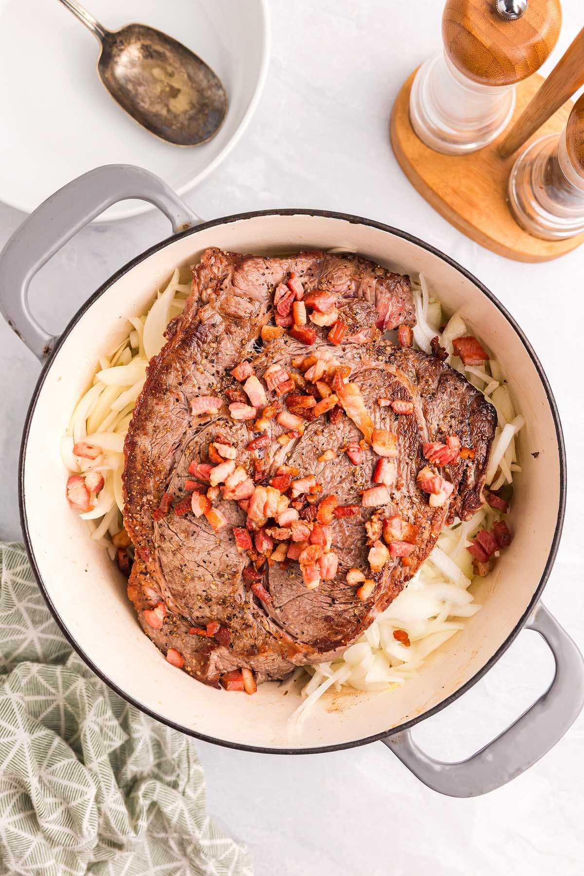 A browned roast topped with chopped bacon sits on sliced onions in a white pot, evoking the rich flavors of pasta alla Genovese, next to seasonings and a spoon.