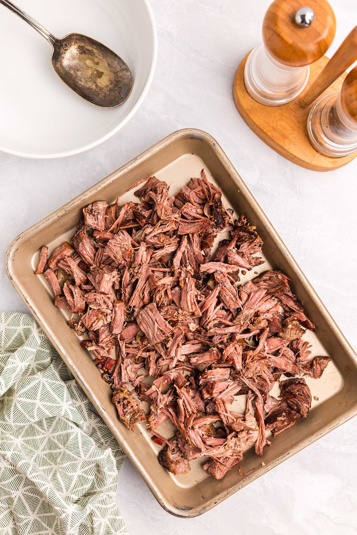 Shredded roast beef on a baking sheet next to a bowl, spoon, salt and pepper shakers, and a green towel&mdash;perfect for preparing pasta alla Genovese.