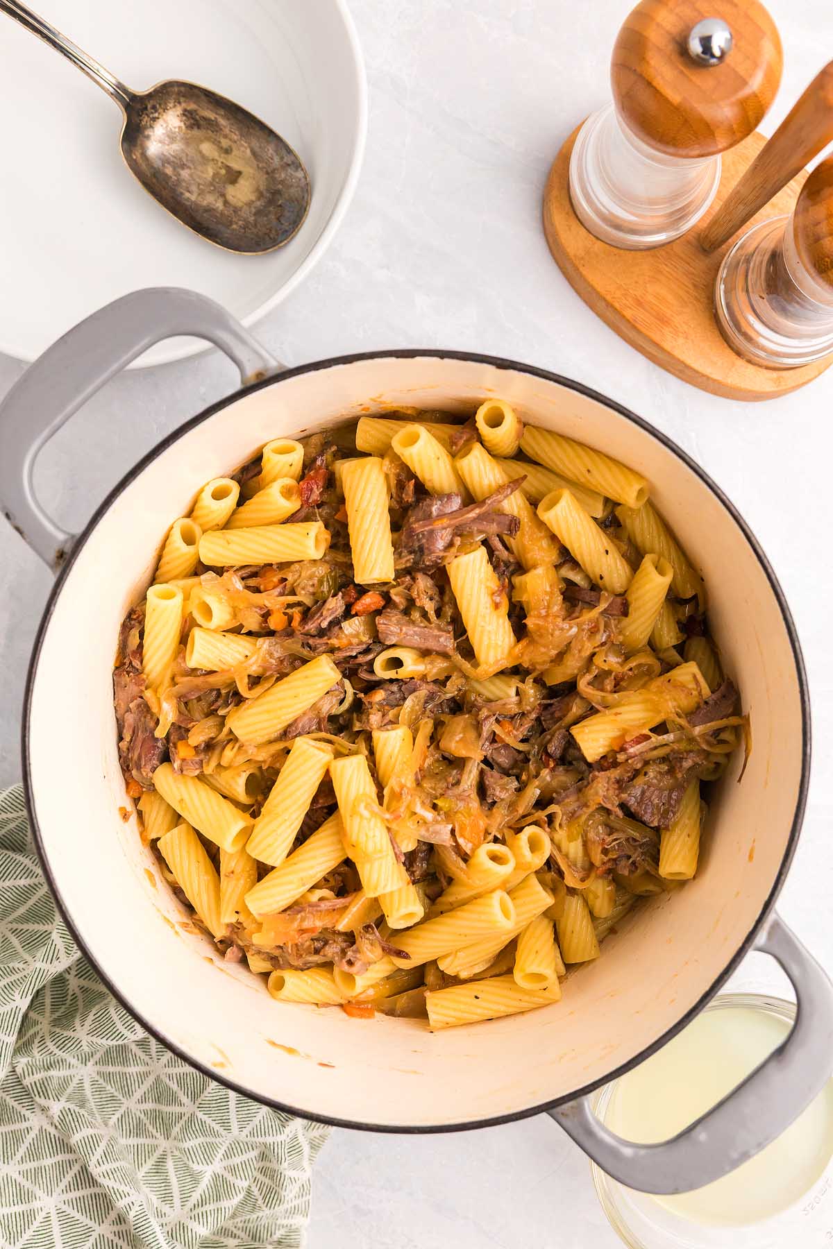 Rigatoni pasta alla Genovese with shredded beef and onions in a white pot, set beside a spoon, seasoning set, and a green towel.
