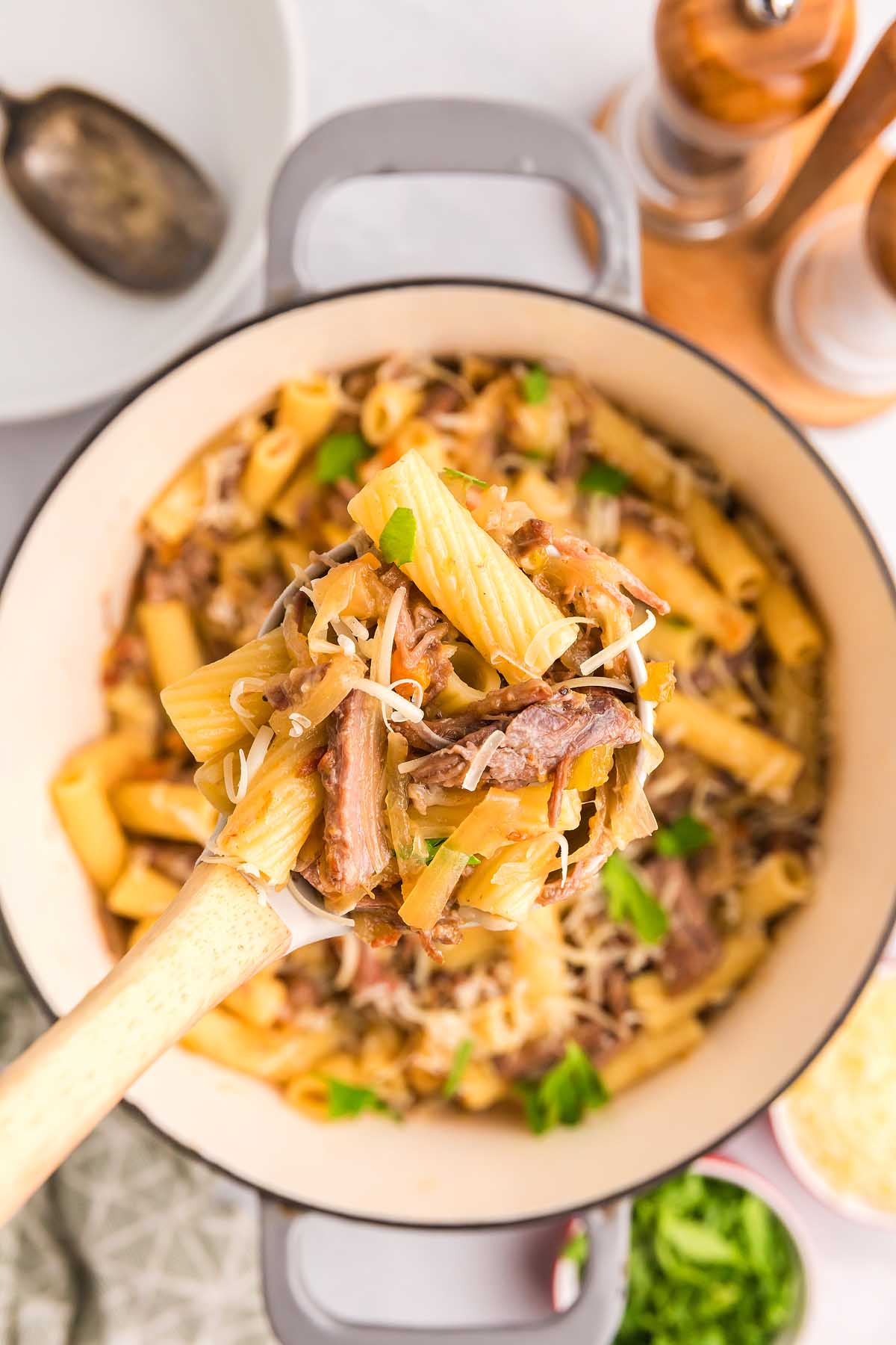 A wooden spoon lifts cheesy rigatoni pasta alla Genovese with shredded meat and herbs from a large pot.
