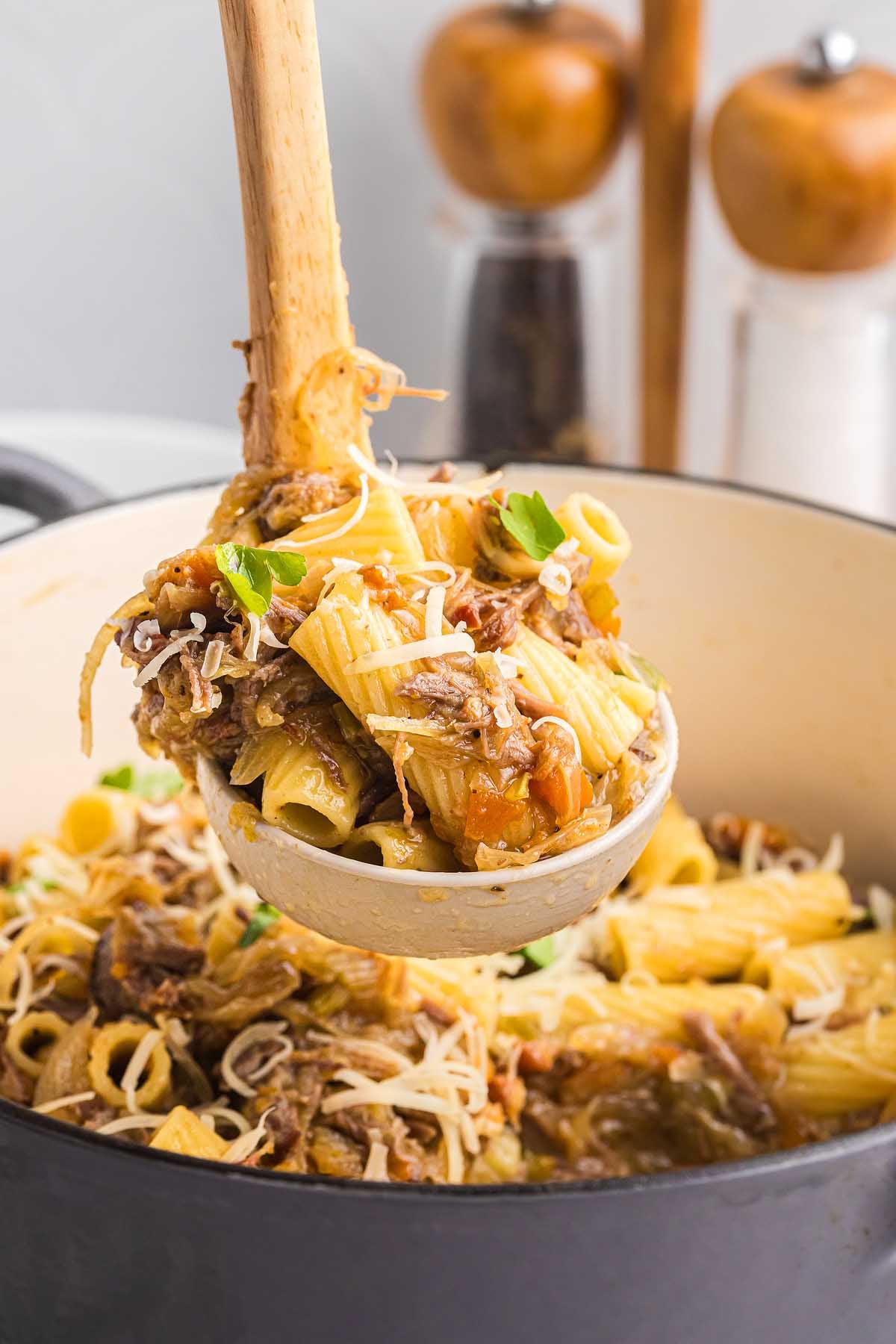 A ladle scoops hearty pasta alla Genovese rigatoni with shredded meat, sauce, and cheese from a pot, with pepper mills in the background.