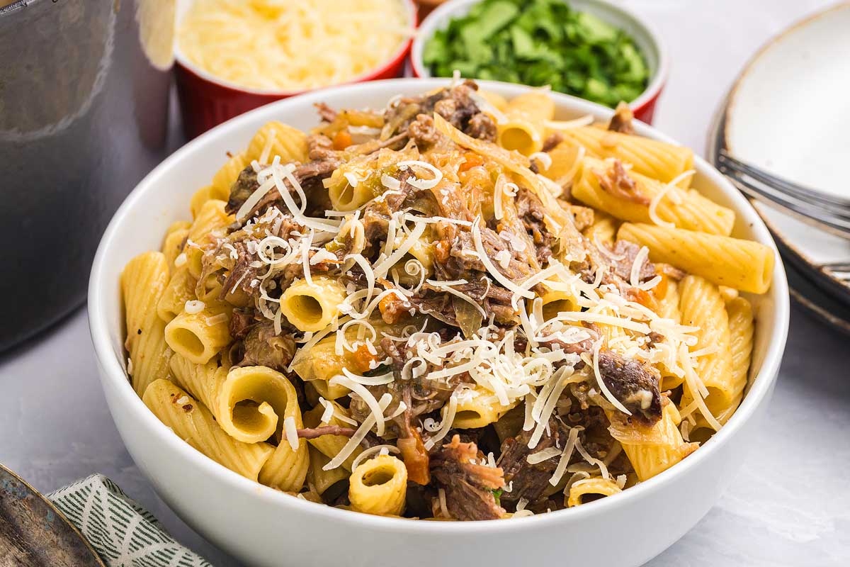 Rigatoni pasta alla Genovese topped with shredded meat and grated cheese, with bowls of cheese and herbs in the background.