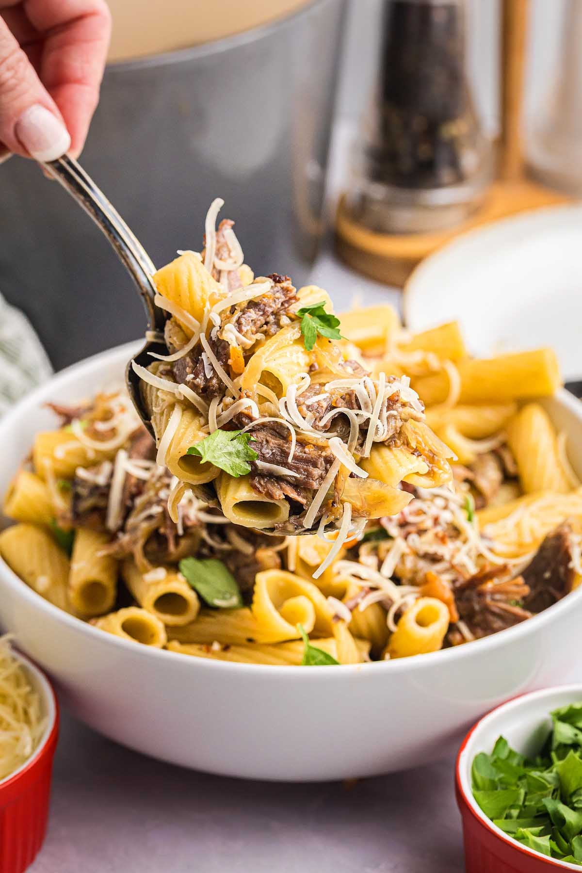 A hand serves cheesy pasta alla Genovese with mushrooms and herbs from a white bowl using a fork.