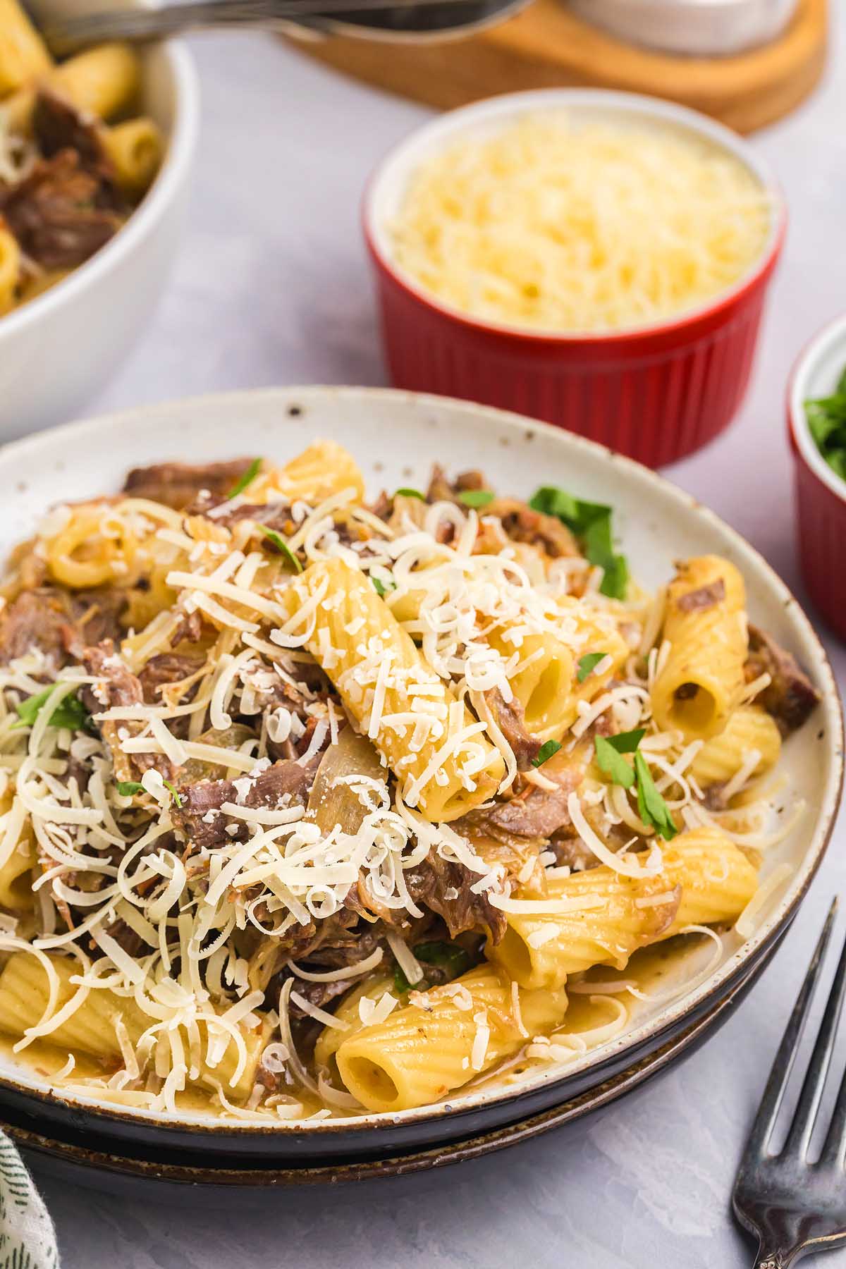 Plate of rigatoni pasta alla Genovese topped with shredded beef, fresh herbs, and grated cheese on a table.
