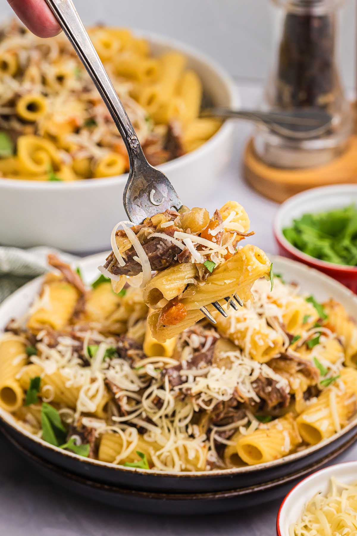 A fork holds cheesy rigatoni pasta alla Genovese with herbs above a bowl filled with more pasta and shredded cheese.