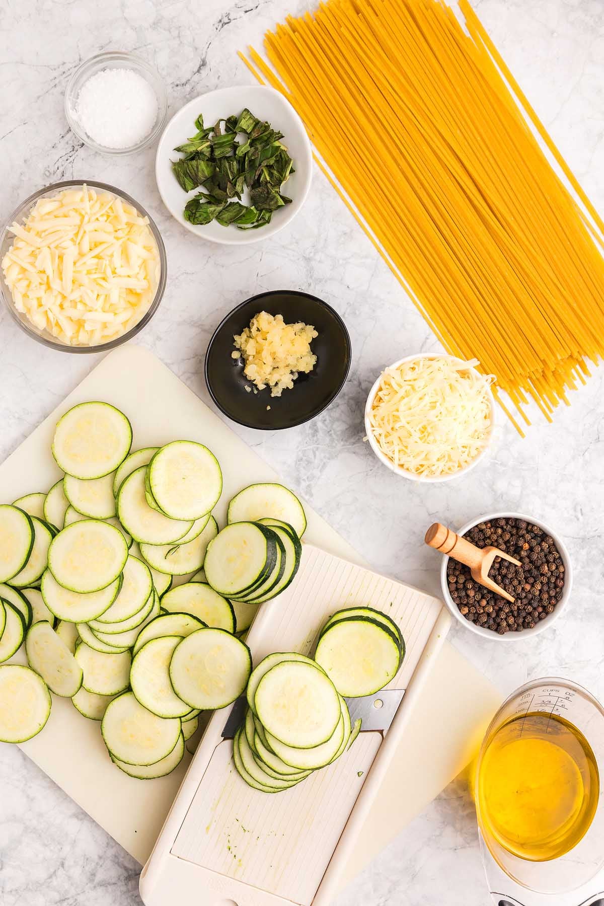 Sliced zucchini, dry spaghetti, cheese, basil, garlic, oil, salt, and pepper arranged on a marble countertop&mdash;perfect ingredients for pasta alla Nerano.