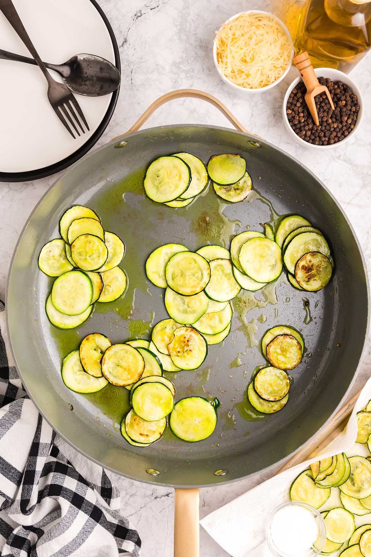 Sliced zucchini saut&eacute;ing in a skillet, inspired by pasta alla Nerano, with cheese, pepper, and utensils nearby on a marble surface.