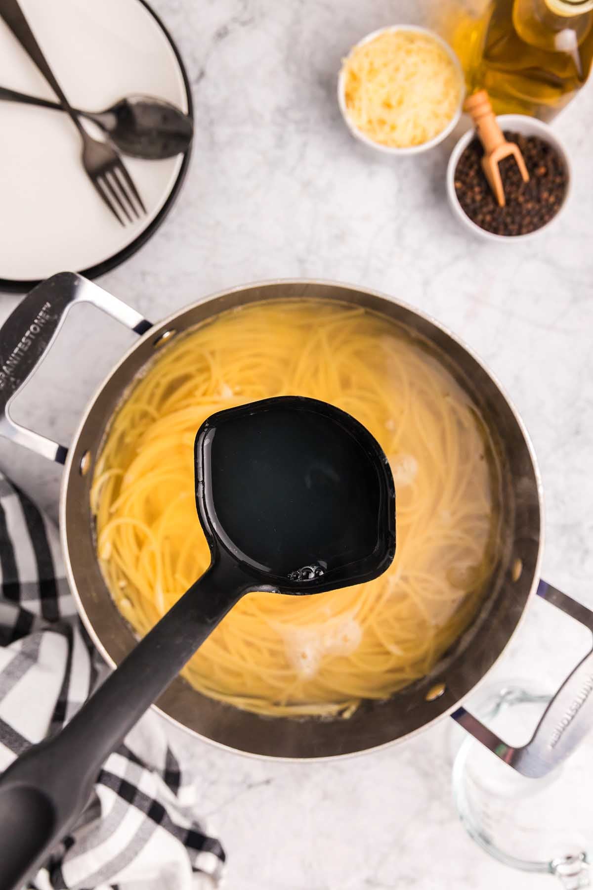 A ladle lifts starchy pasta water from a pot of cooking spaghetti, a key step in preparing pasta alla Nerano, on a kitchen counter.
