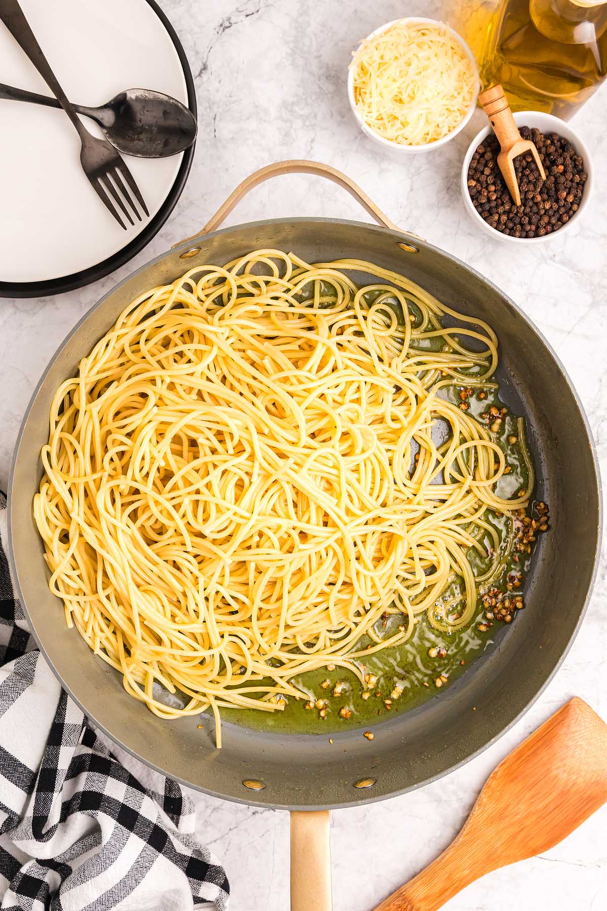 Cooked spaghetti pasta alla Nerano in a large skillet with olive oil, pepper, cheese, and utensils on a marble countertop.