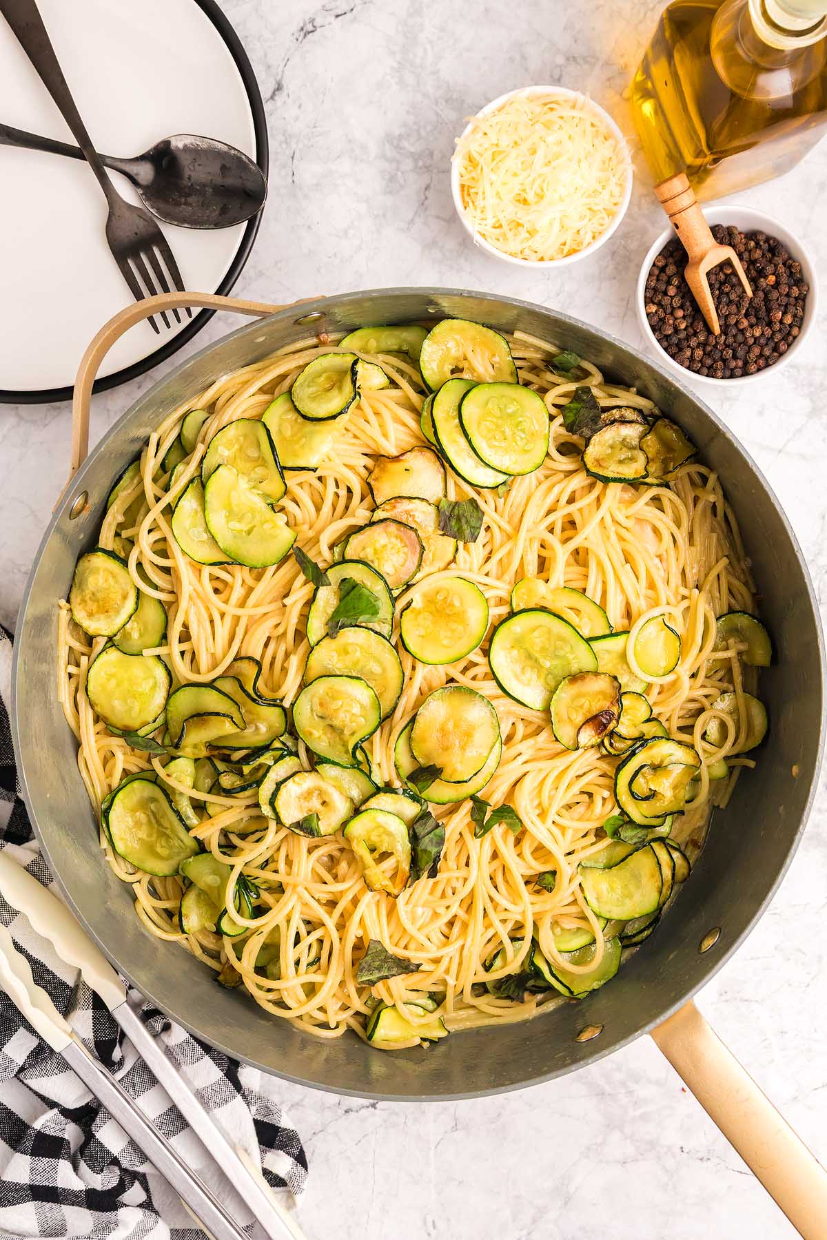 Pasta alla Nerano with saut&eacute;ed zucchini in a large pan, surrounded by cheese, black pepper, oil, and utensils on a marble surface.