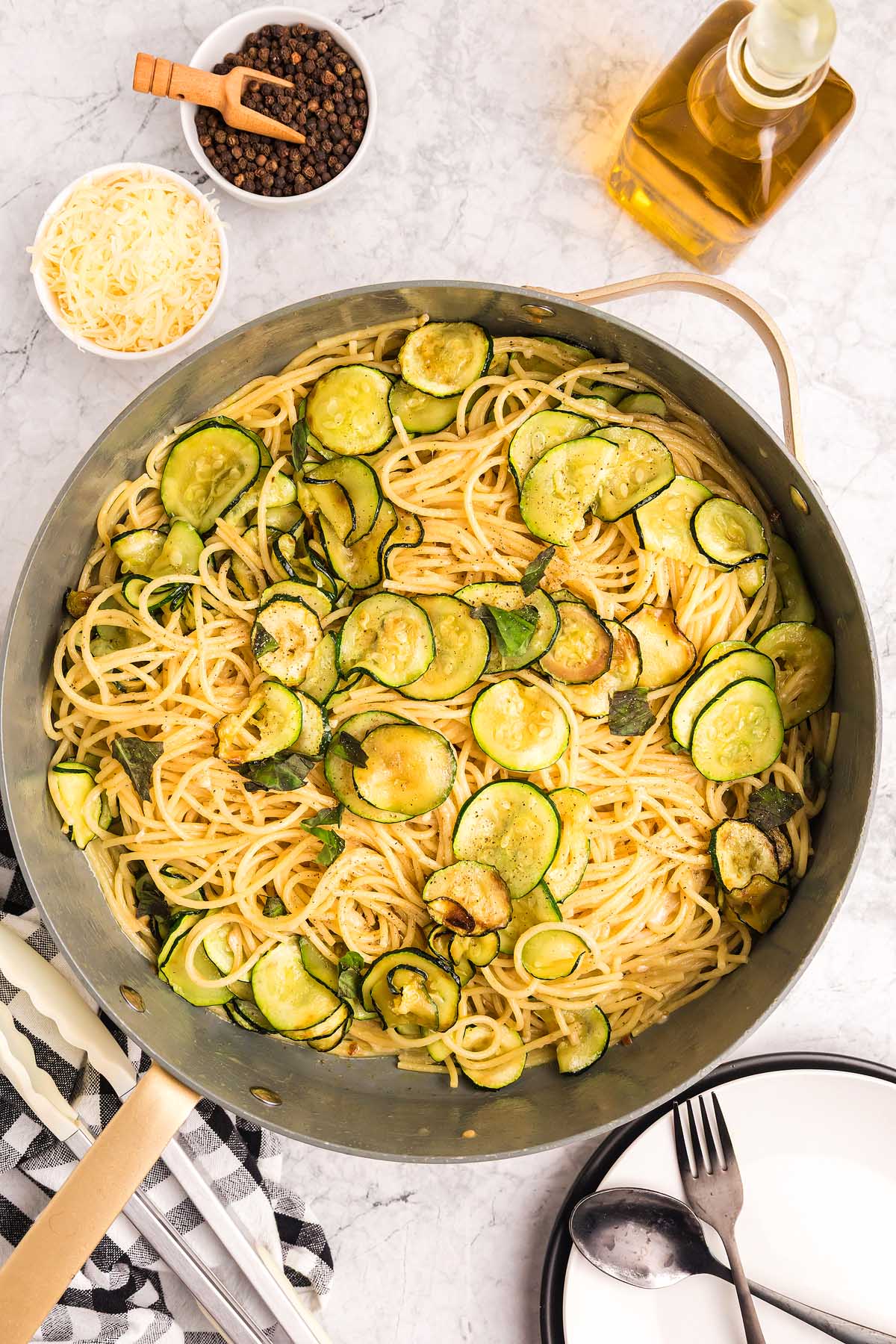 A pan of pasta alla Nerano with saut&eacute;ed zucchini slices, surrounded by grated cheese, pepper, and olive oil on a marble surface.