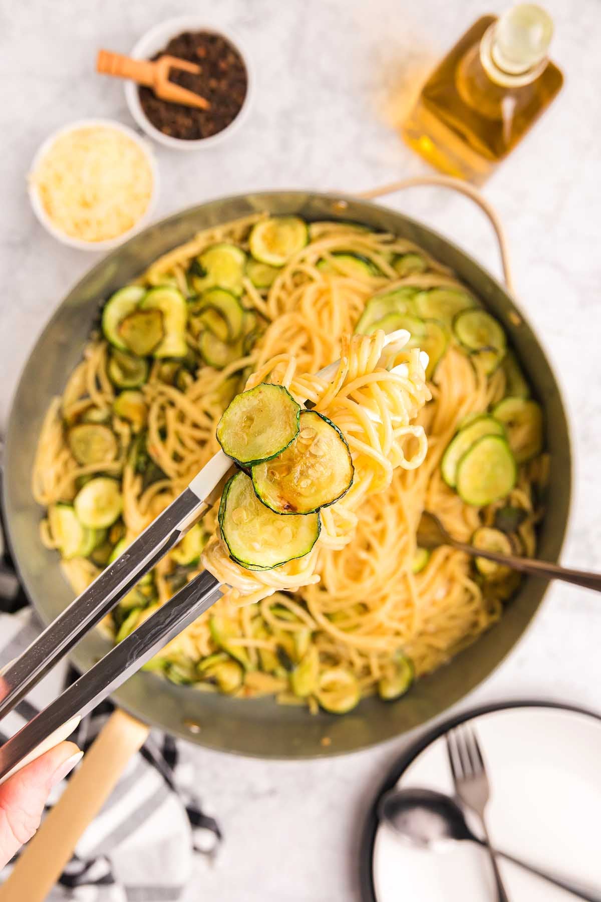 Tongs lifting pasta alla Nerano with saut&eacute;ed zucchini from a pan, surrounded by ingredients and plates.