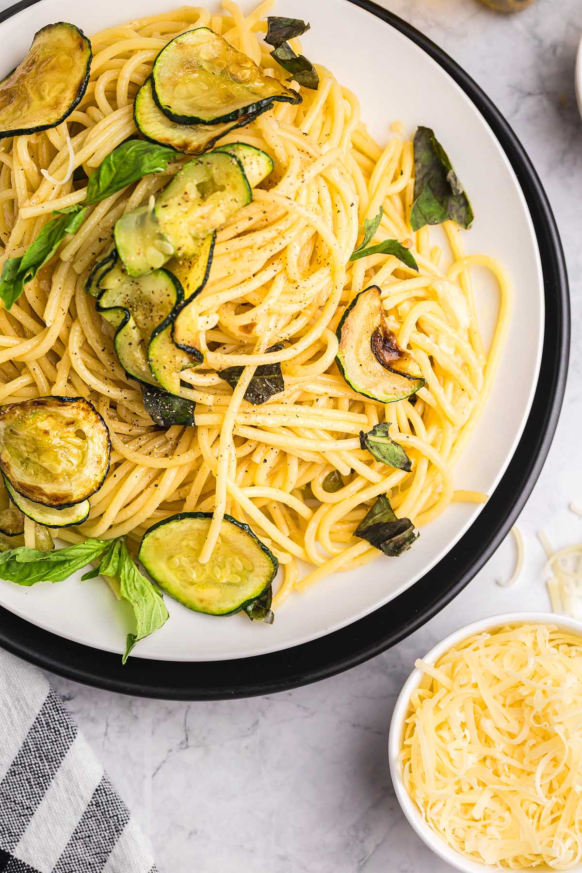 Plate of pasta alla Nerano featuring saut&eacute;ed zucchini and fresh basil, served alongside a bowl of shredded cheese.