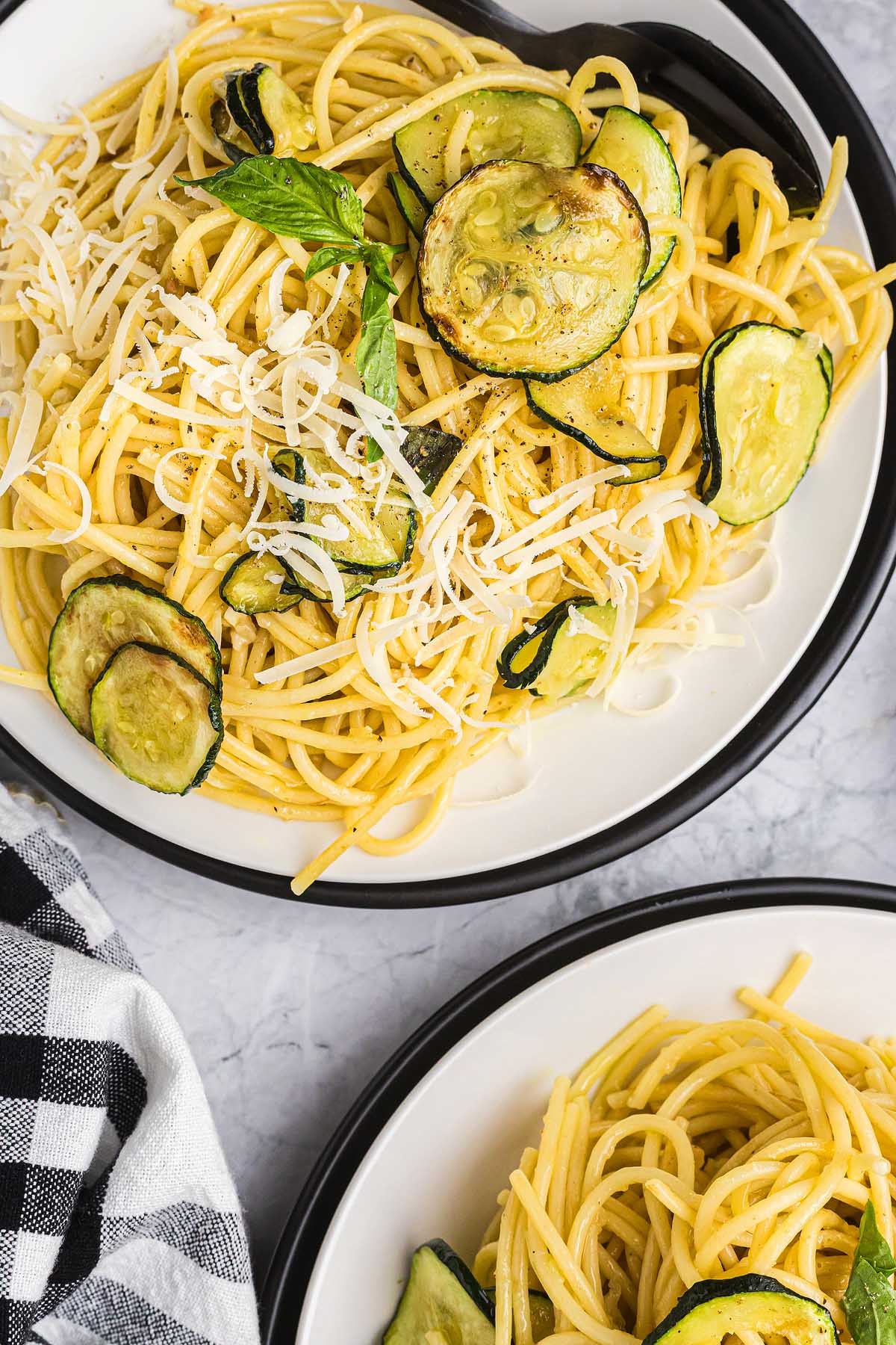Spaghetti pasta alla Nerano with saut&eacute;ed zucchini and shredded cheese, served on white plates with a black-and-white napkin.